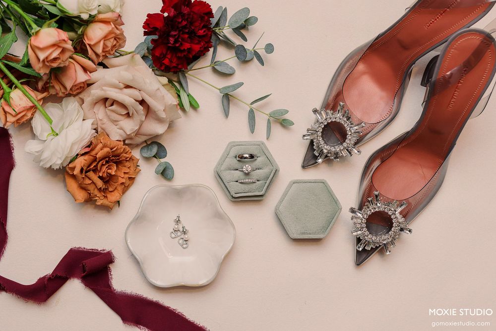 Wedding details flatlay with deep red roses, vintage rings, and delicate blush ribbons on a soft pink background.