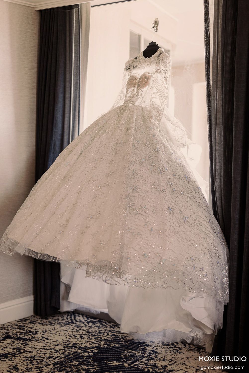 An elegant white ball gown wedding dress with intricate lace details hangs on display against dark curtains.