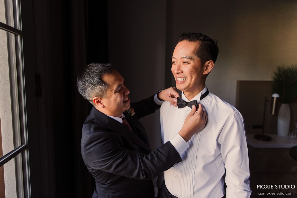 Wedding preparation scene with two people helping adjust formal attire in natural window light.