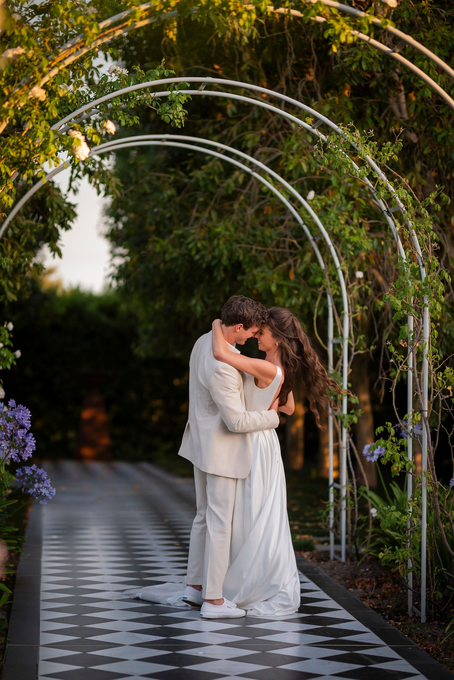 A couple embraces on a black and white checkered walkway beneath a white archway in a garden setting at sunset.