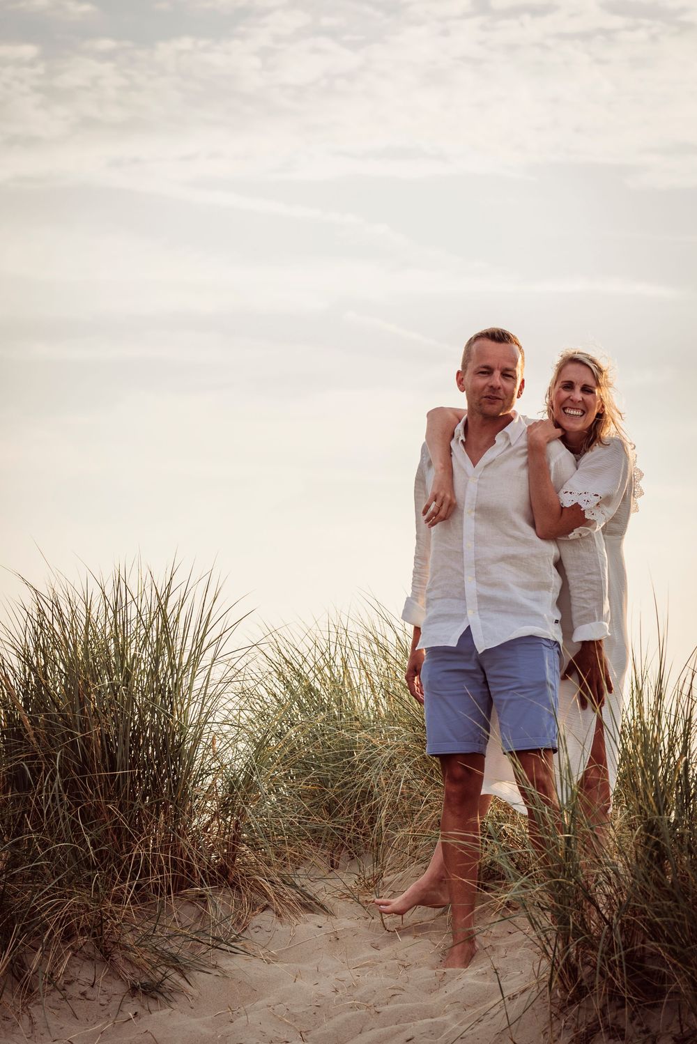 Smiling couple embracing on sandy beach dunes with tall grass, dressed in white casual outfits.