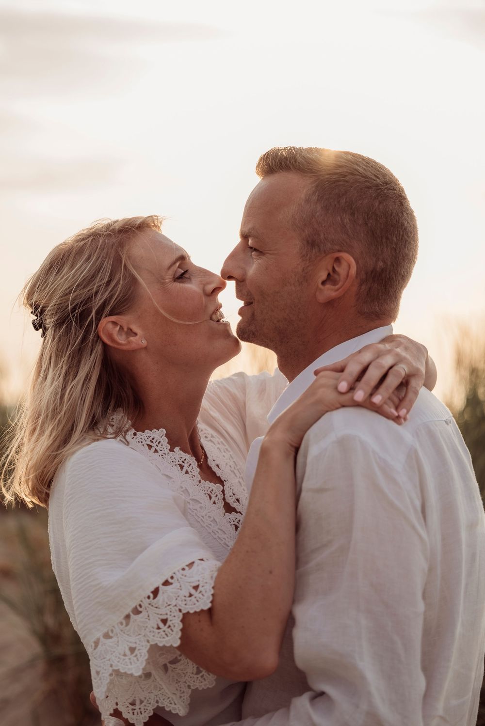 Romantic couple about to kiss at sunset on the beach, both wearing white lace and linen outfits.