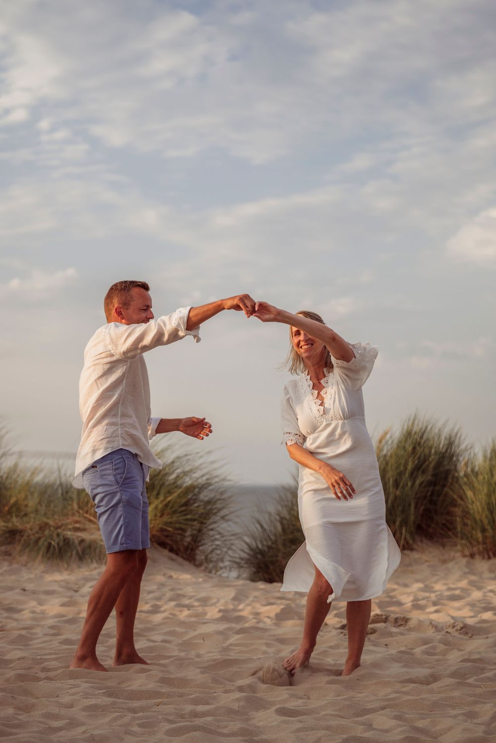 Joyful couple dancing together on sandy beach dunes, holding hands and spinning in white outfits.