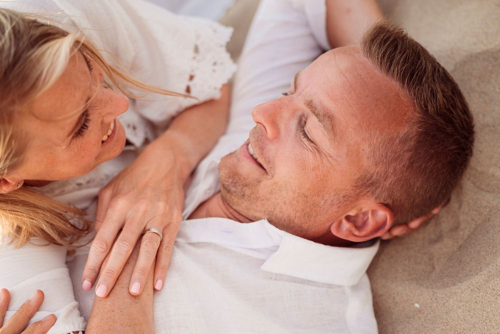 A couple lying on sandy beach, woman gazing lovingly at smiling man wearing white shirts.