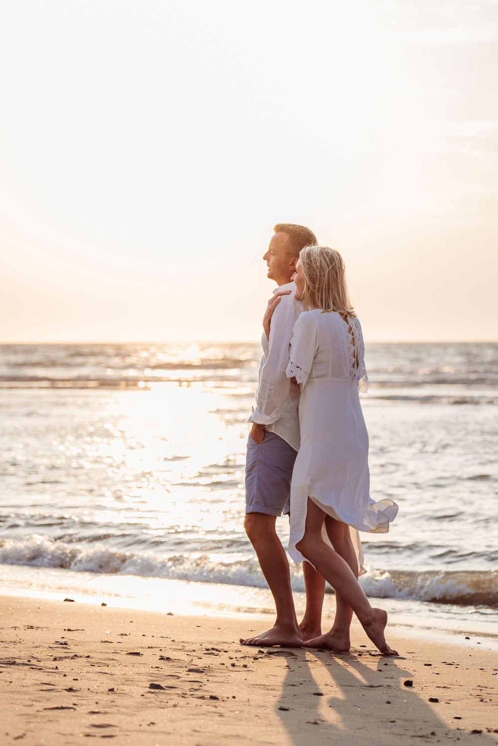 A romantic couple walking barefoot along a sandy beach holding each other at golden sunset.