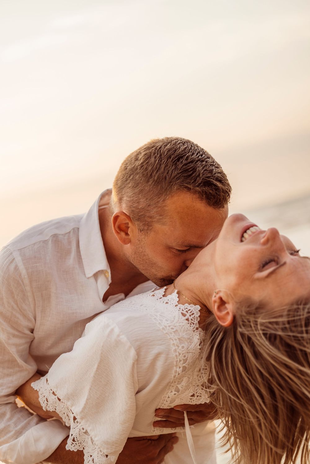 A man kissing a laughing woman on the cheek while holding her on a beach at sunset.