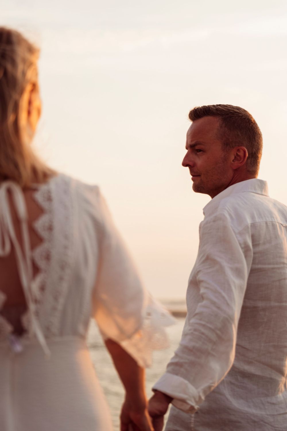 A couple holding hands on a beach at sunset, man gazing at woman in white lace dress.