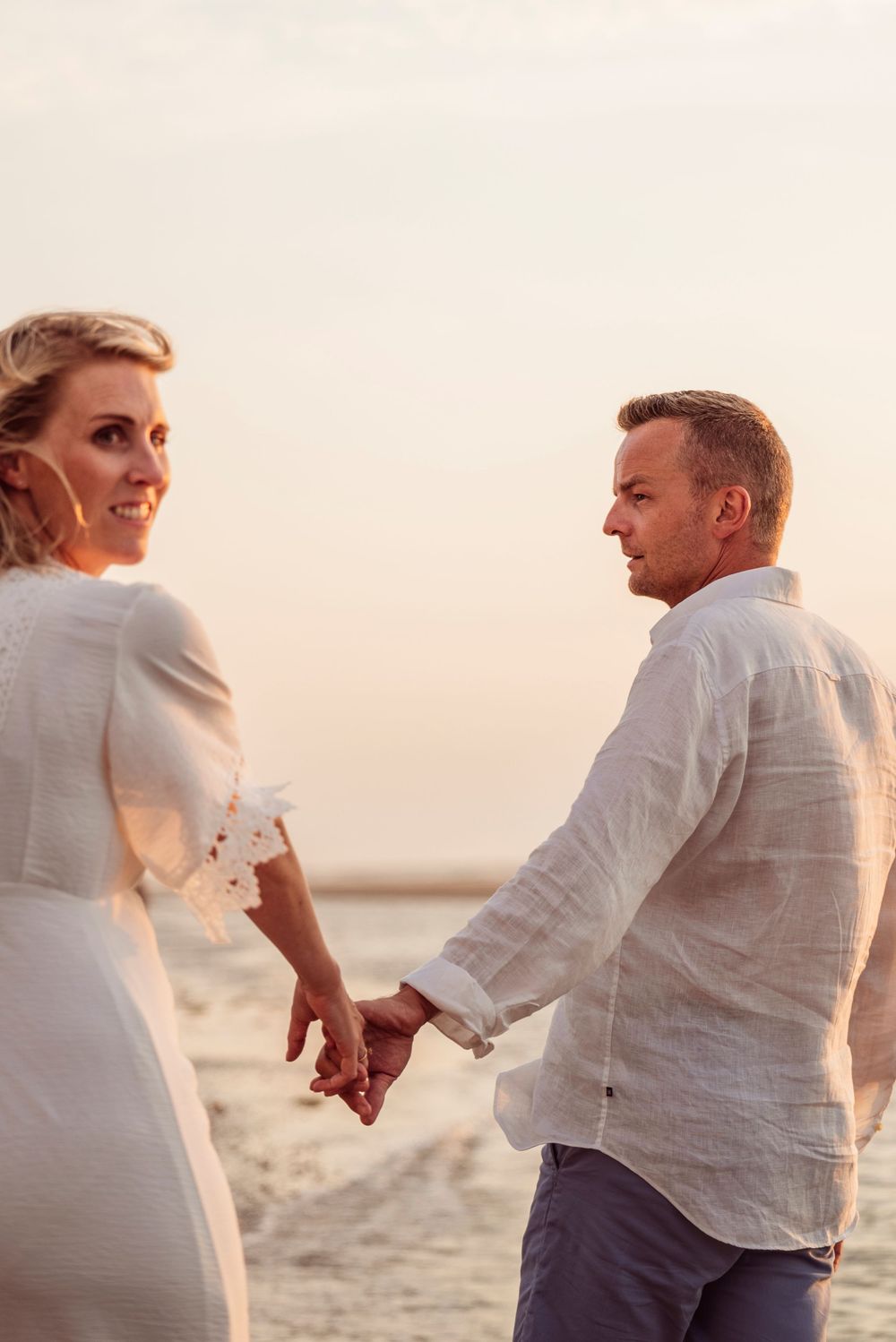 A smiling couple in white outfits holding hands and looking back at each other on a beach.