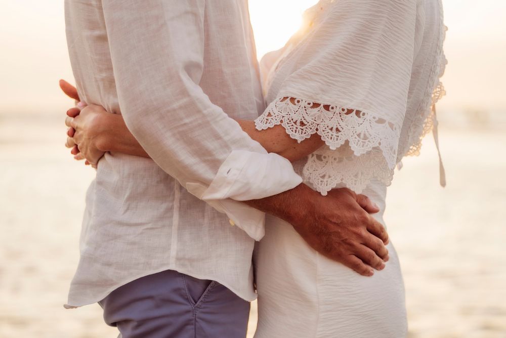 Couple embracing at sunset on the beach, wearing white outfits, close-up of their arms wrapped around each other.