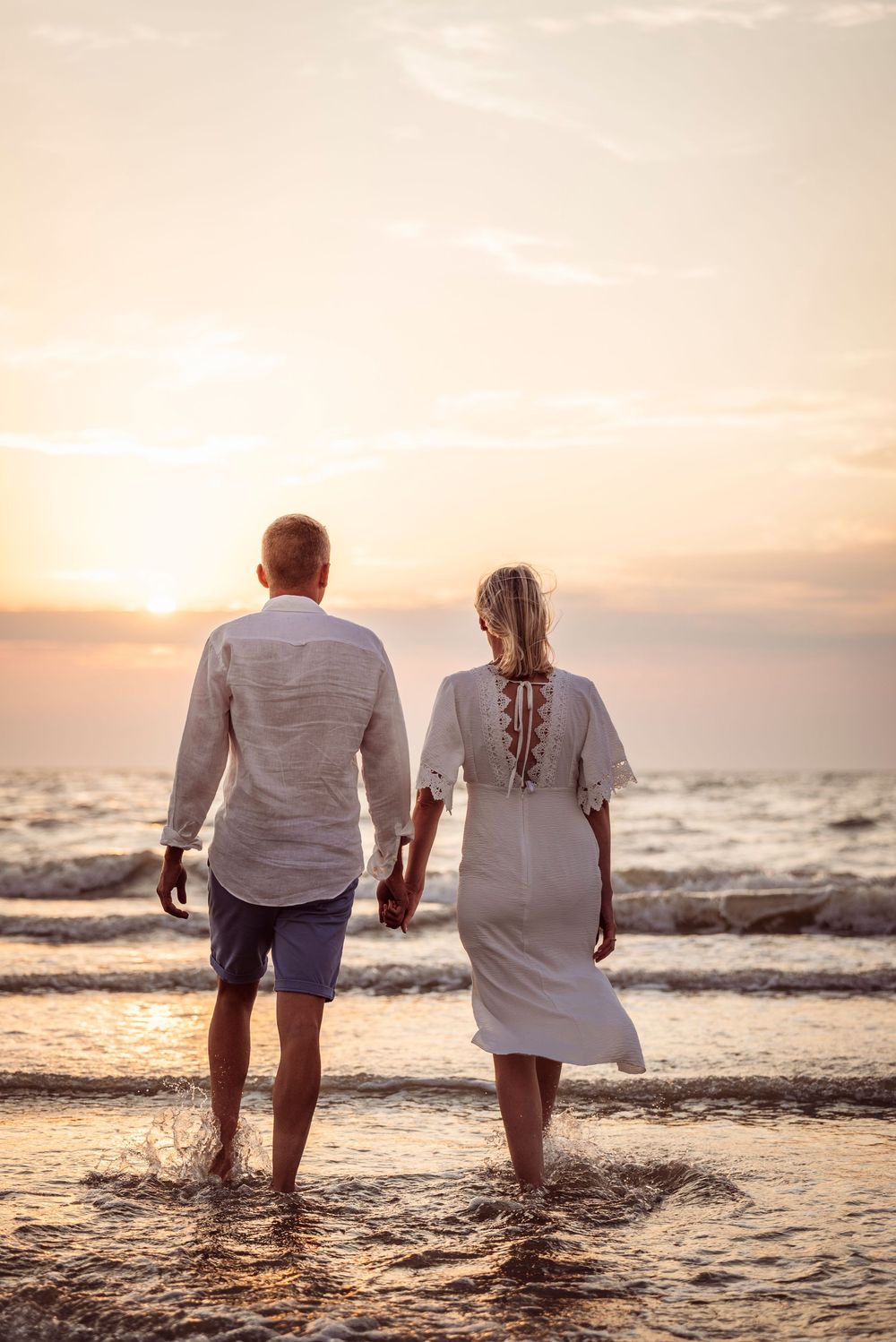Couple holding hands walking toward the ocean at golden sunset, viewed from behind on a beach.