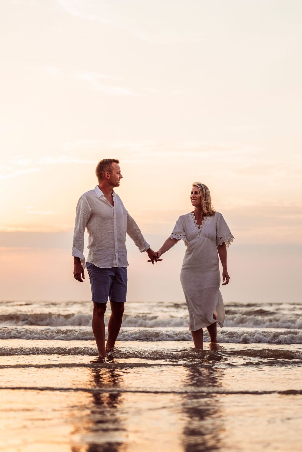 Romantic couple holding hands and walking along the shoreline at sunset, both dressed in white.