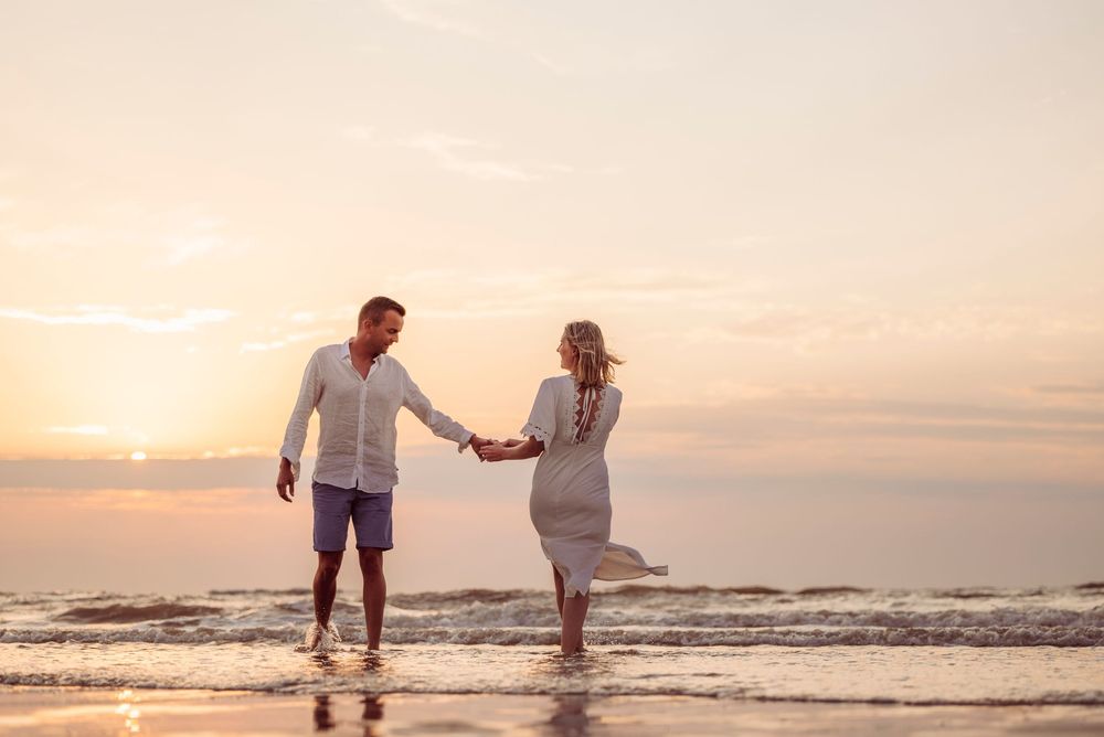 Couple holding hands walking along shoreline at golden sunset, wearing coordinating white outfits.