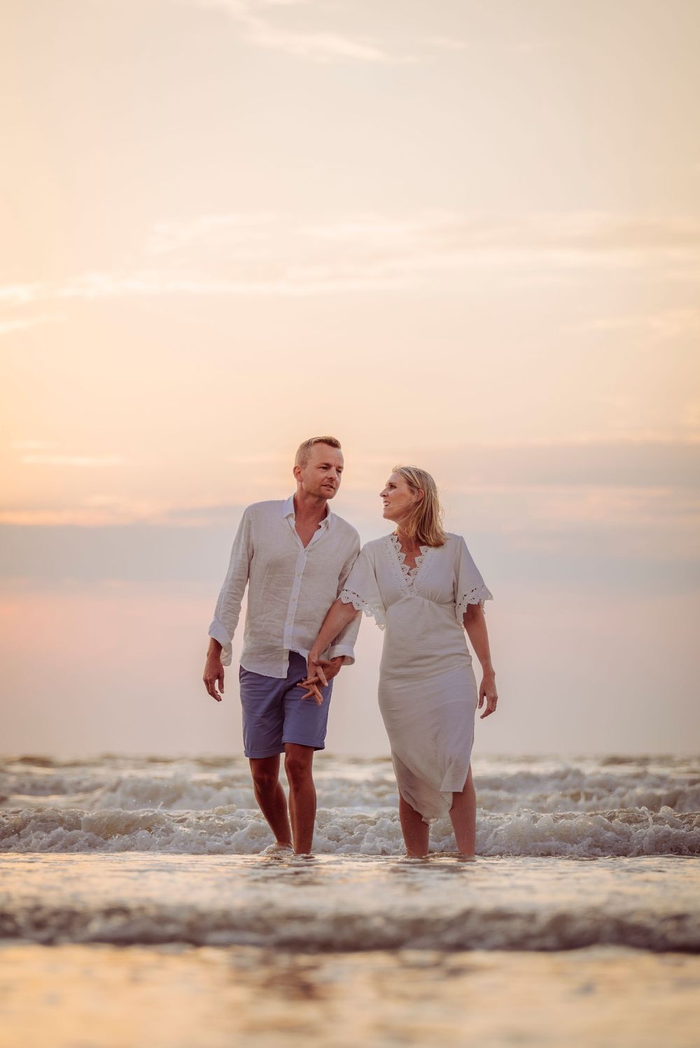 Happy couple walking hand in hand through shallow ocean waves at a beautiful beach sunset.