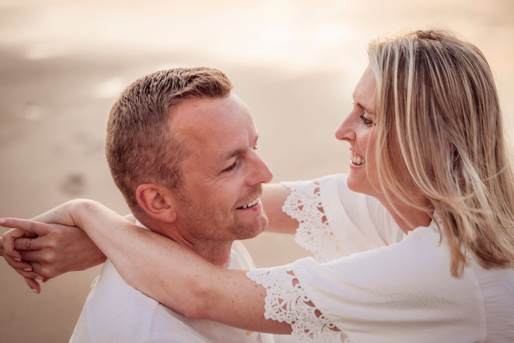 Happy couple smiling at each other outdoors, woman in white lace top embracing mans shoulders.