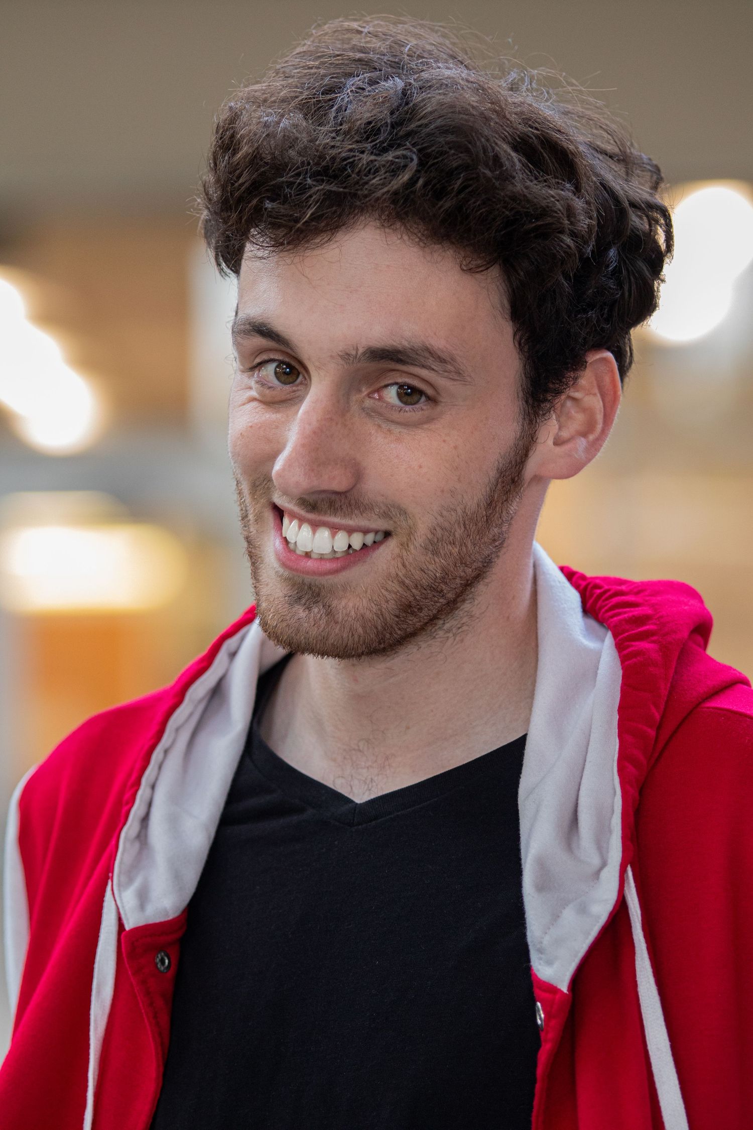 A young person wearing a red hoodie and black v-neck shirt smiles warmly at the camera.