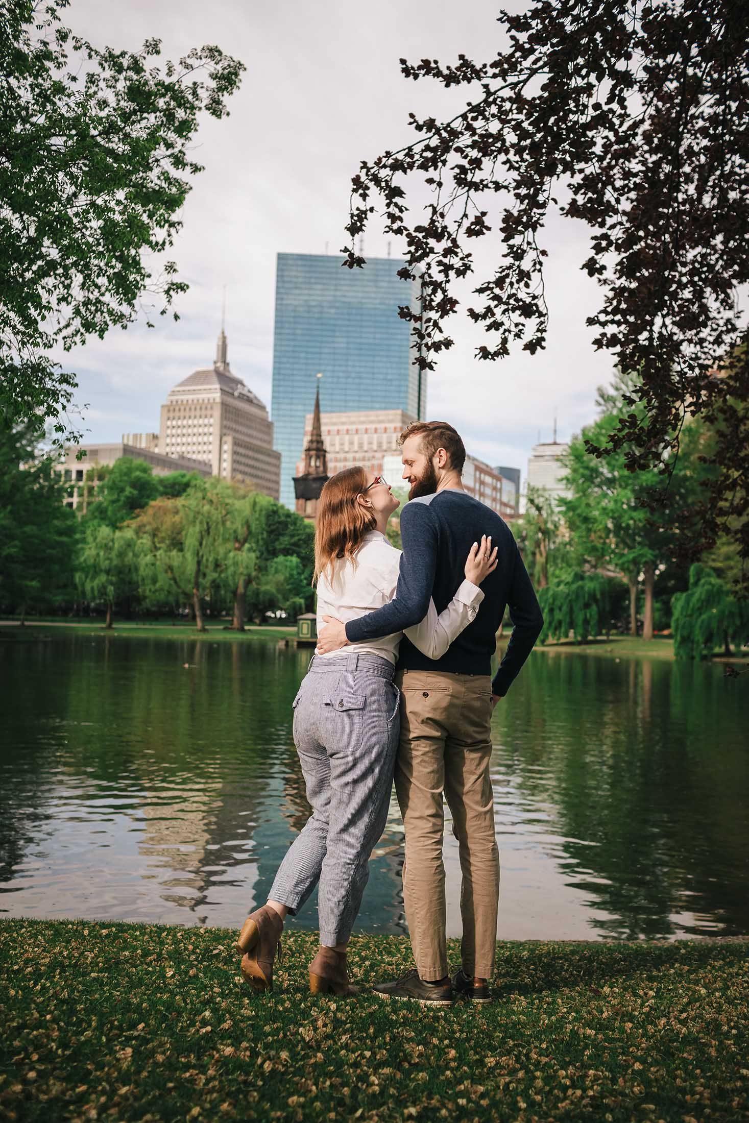 Engaged couple admiring Boston skyline and lagoon reflections during engagement session in Boston Public Garden