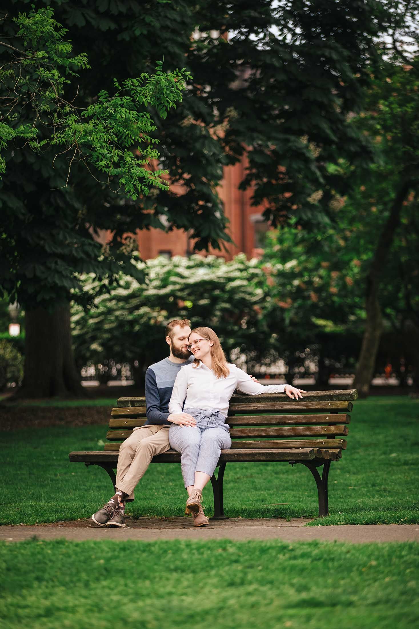 Romantic portrait of engaged couple sitting together on park bench in Boston Public Garden near George Robert White Memorial