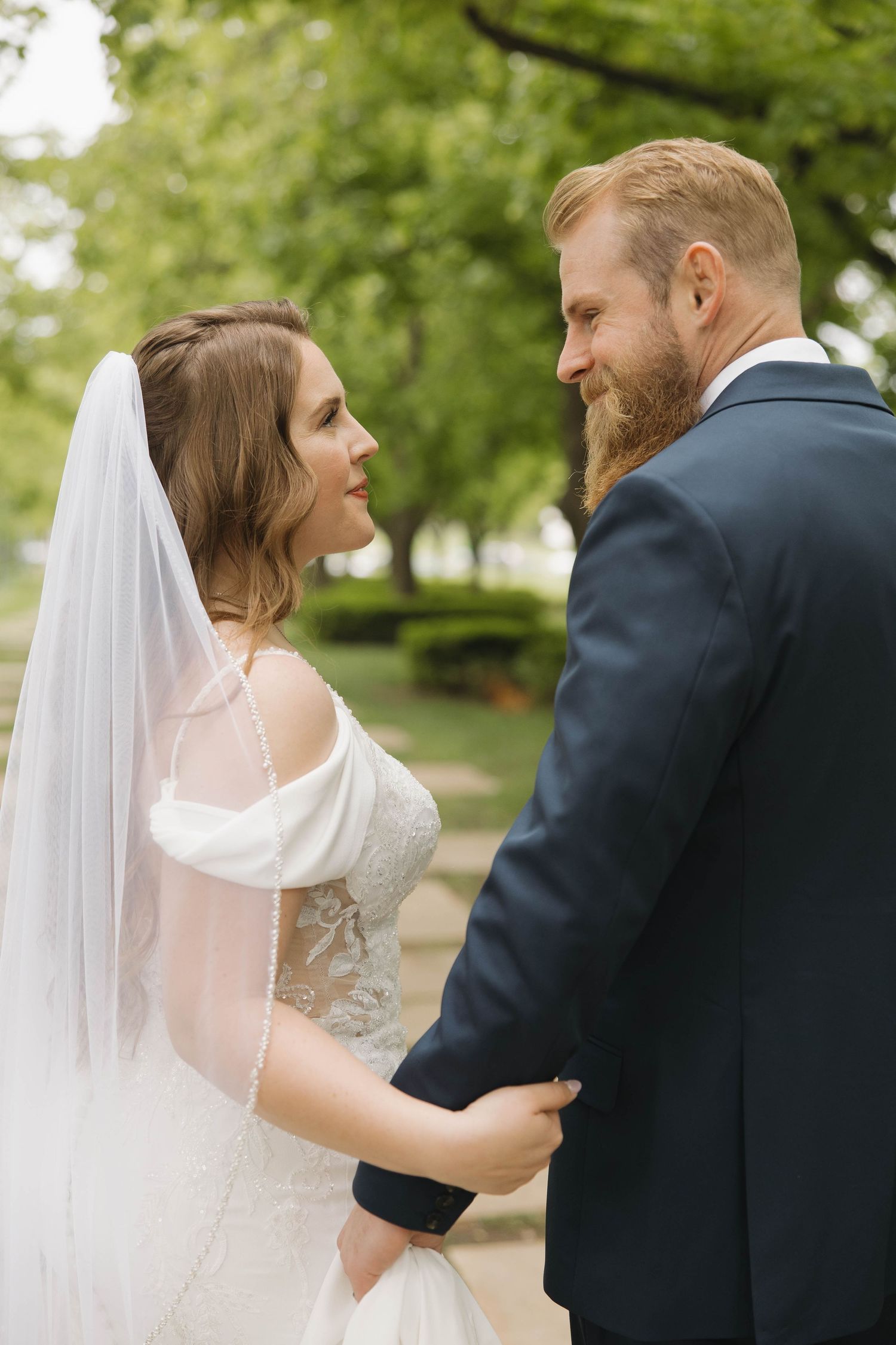 A bride in a white gown with flowing veil and groom in navy suit share an intimate moment outdoors among green trees.