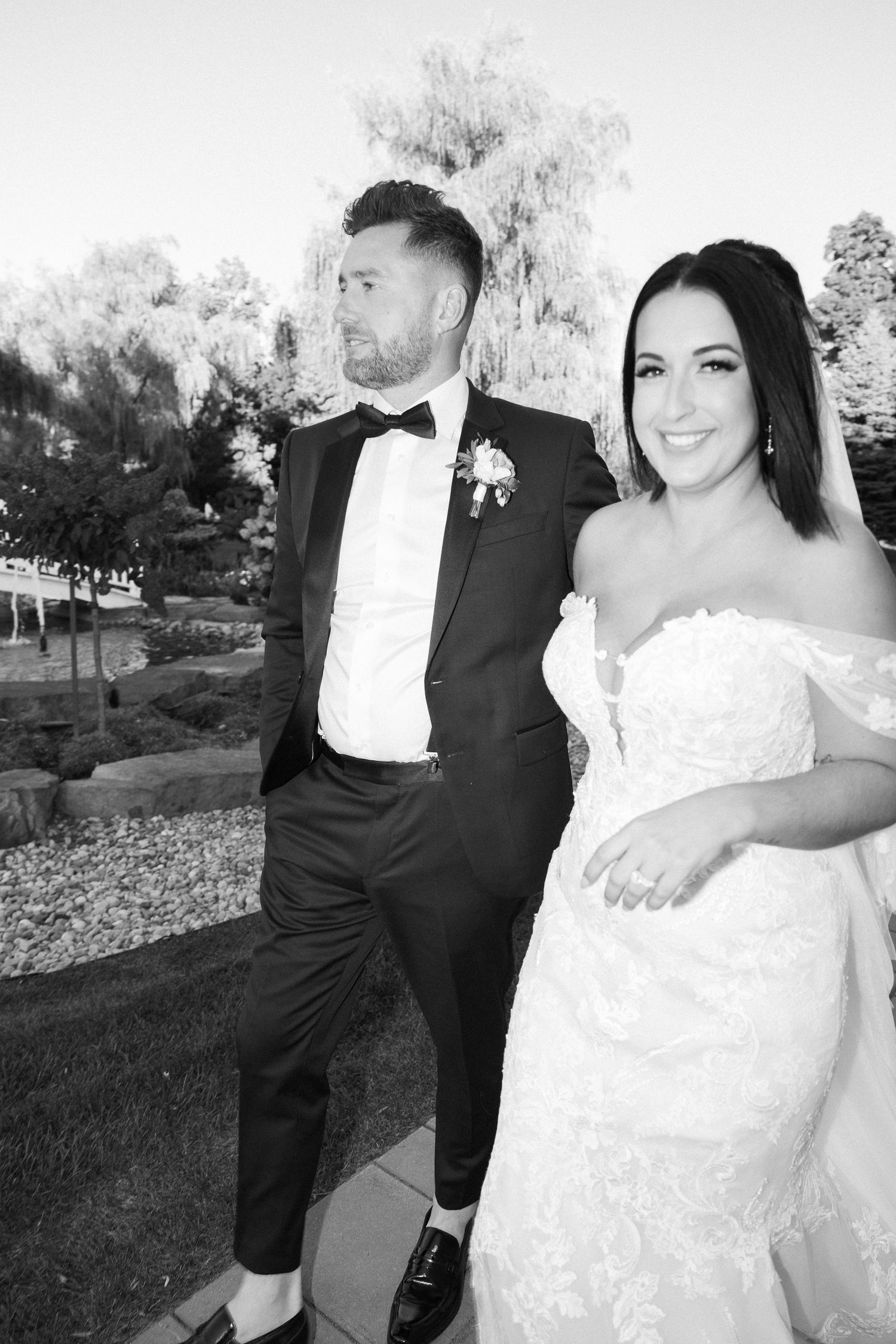 Groom in black tuxedo kisses brides temple as she holds lace ball gown skirt at sunset.