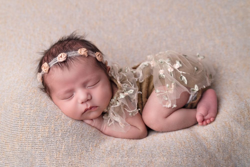 Sleeping newborn baby girl in beige lace romper and floral headband on soft tan blanket.