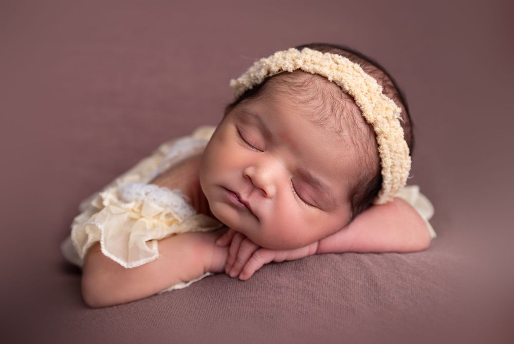 Sleeping newborn baby girl wearing a cream lace headband and outfit, posed on a mauve background.