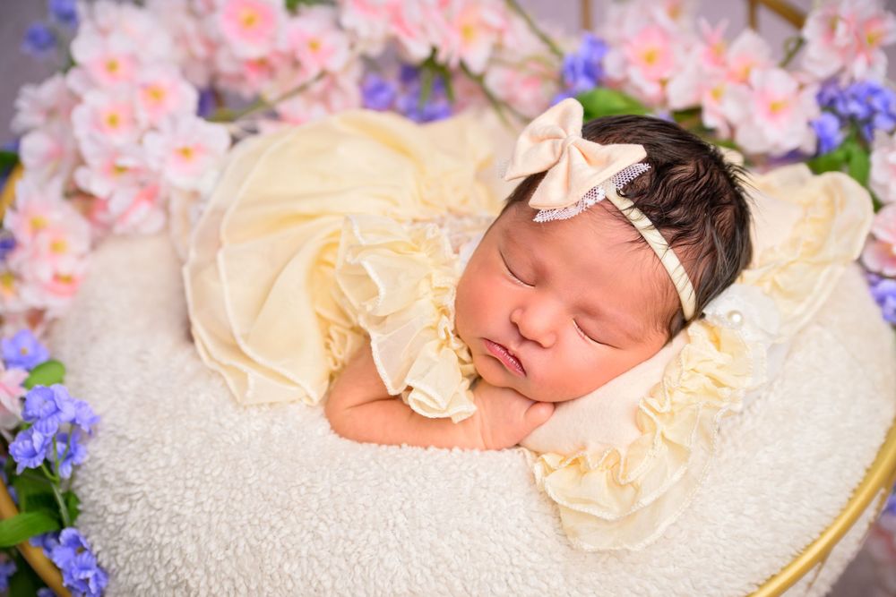 Sleeping newborn baby girl in yellow dress and bow headband nestled in floral arrangement for professional photo.