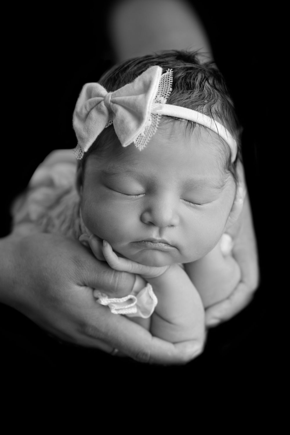 Black and white newborn baby girl sleeping peacefully in parents hands wearing a bow headband.