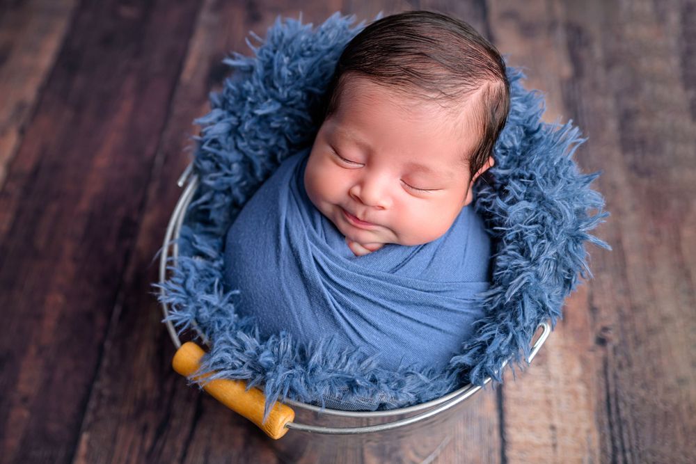 Newborn baby wrapped in blue swaddle sleeping peacefully in a silver bucket with blue fur lining on wood floor.
