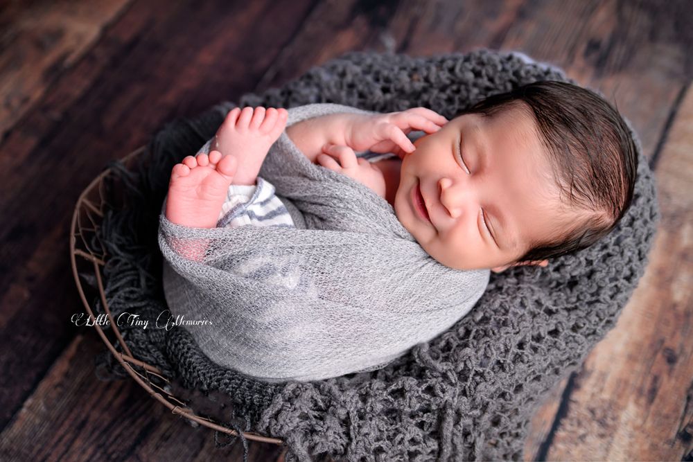 Smiling newborn baby wrapped in gray knit swaddle sleeping peacefully in a rustic wire basket on wood background.