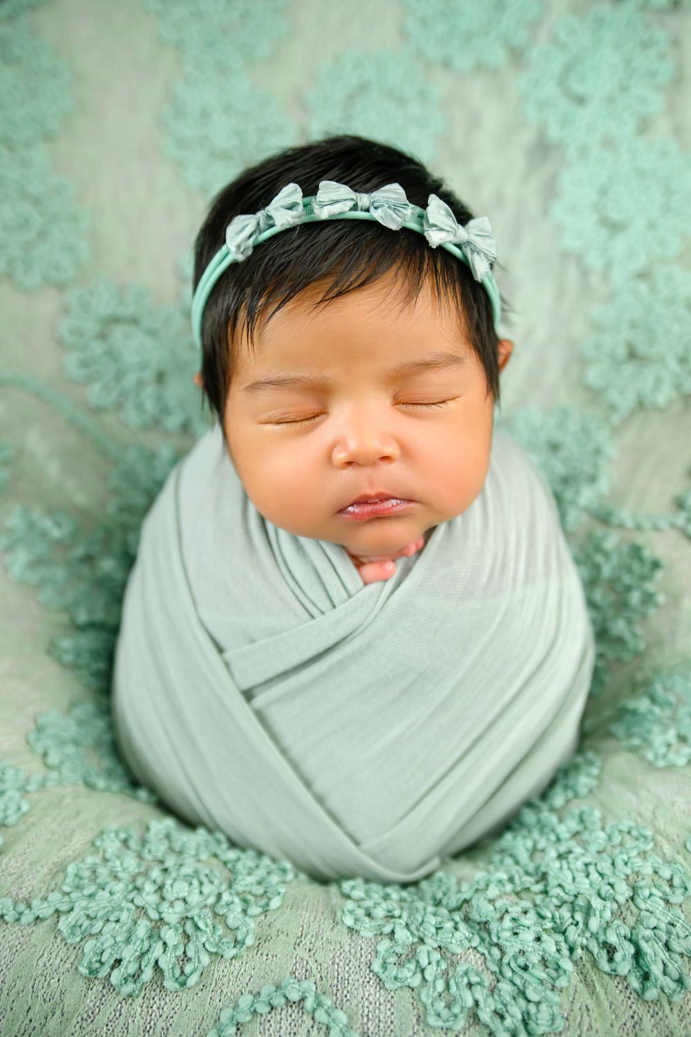 Newborn baby girl swaddled in teal wrap with floral headband, sleeping peacefully on matching lace backdrop.