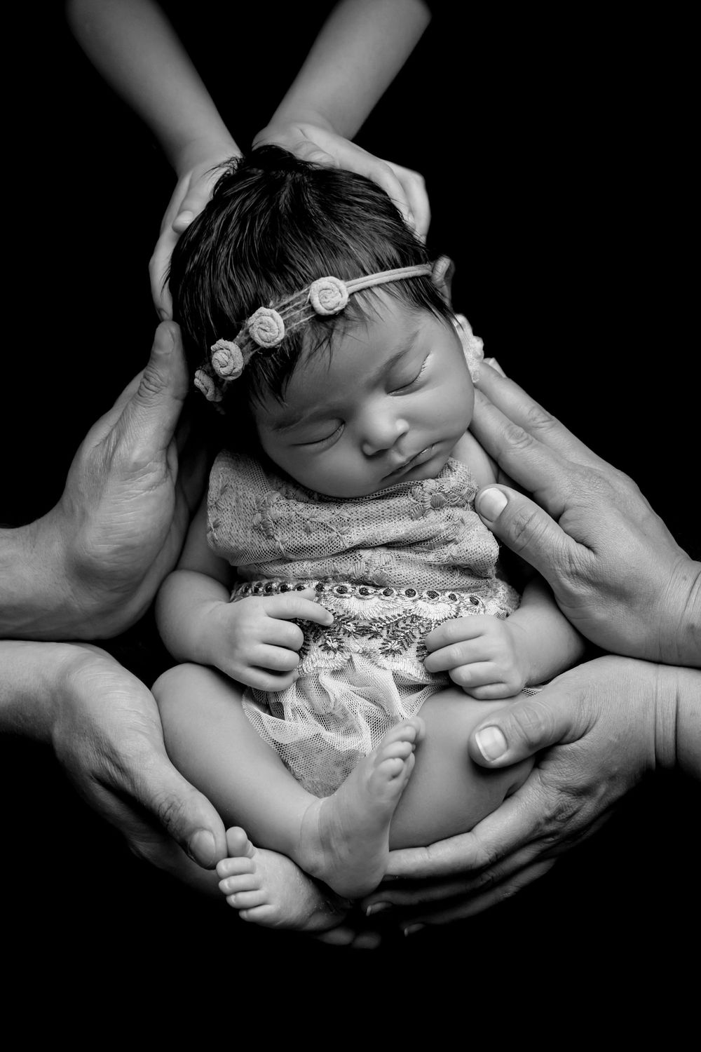 Black and white photo of sleeping newborn baby girl in floral headband cradled by multiple loving hands.