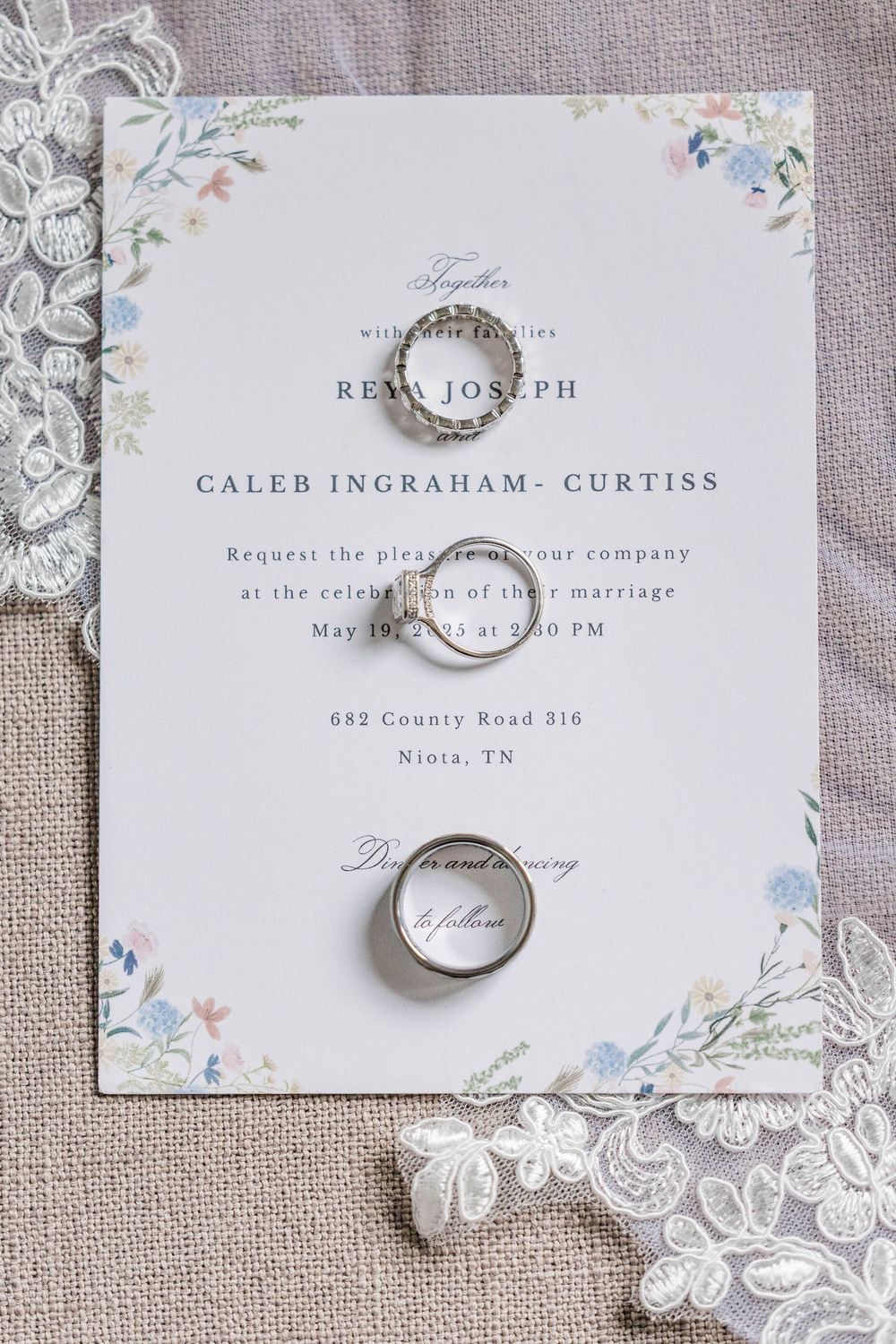 Elegant wedding invitation with floral details and wedding rings on lace background.