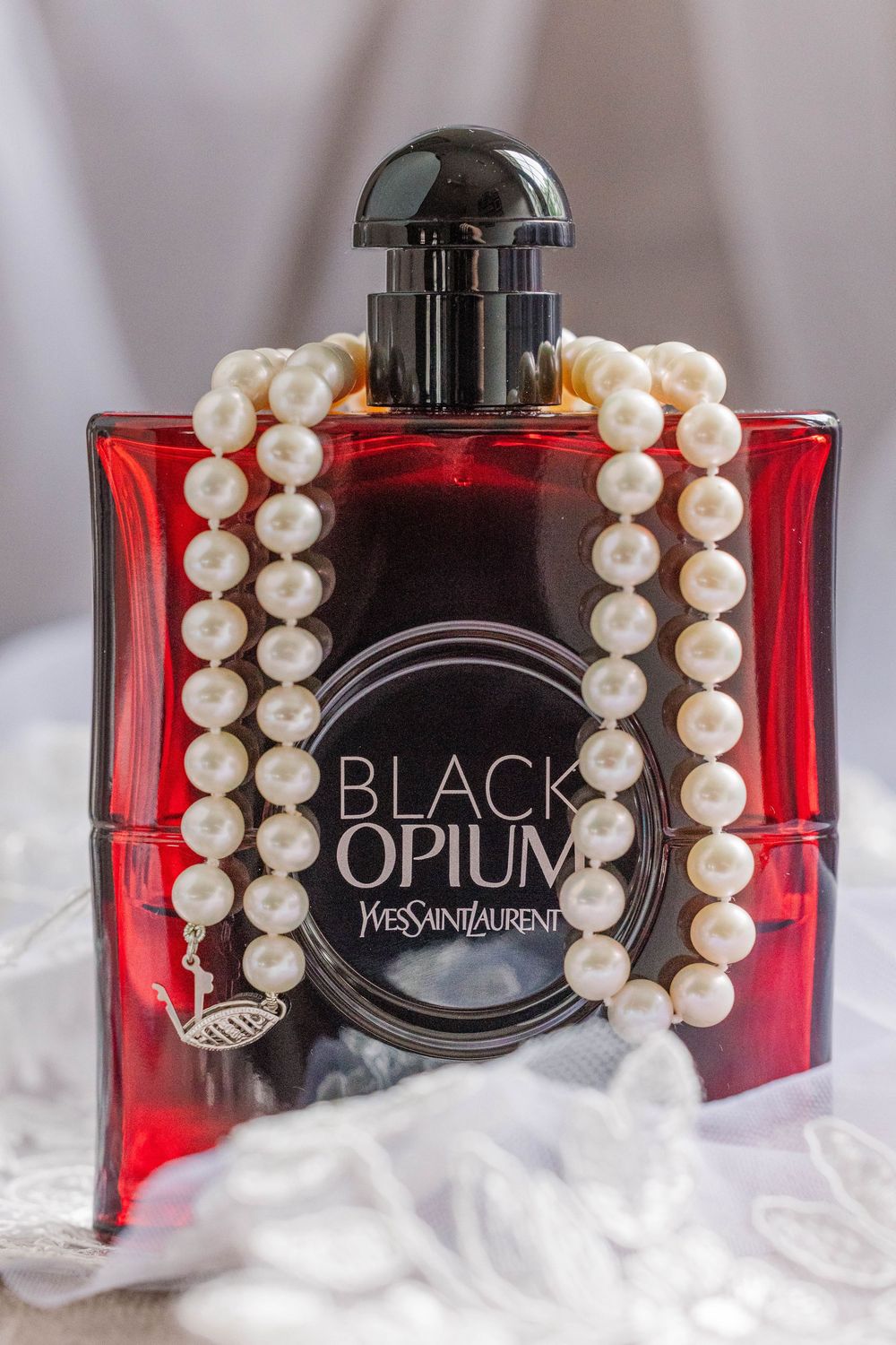 Black Opium perfume bottle by Yves Saint Laurent adorned with a pearl necklace against a white background.