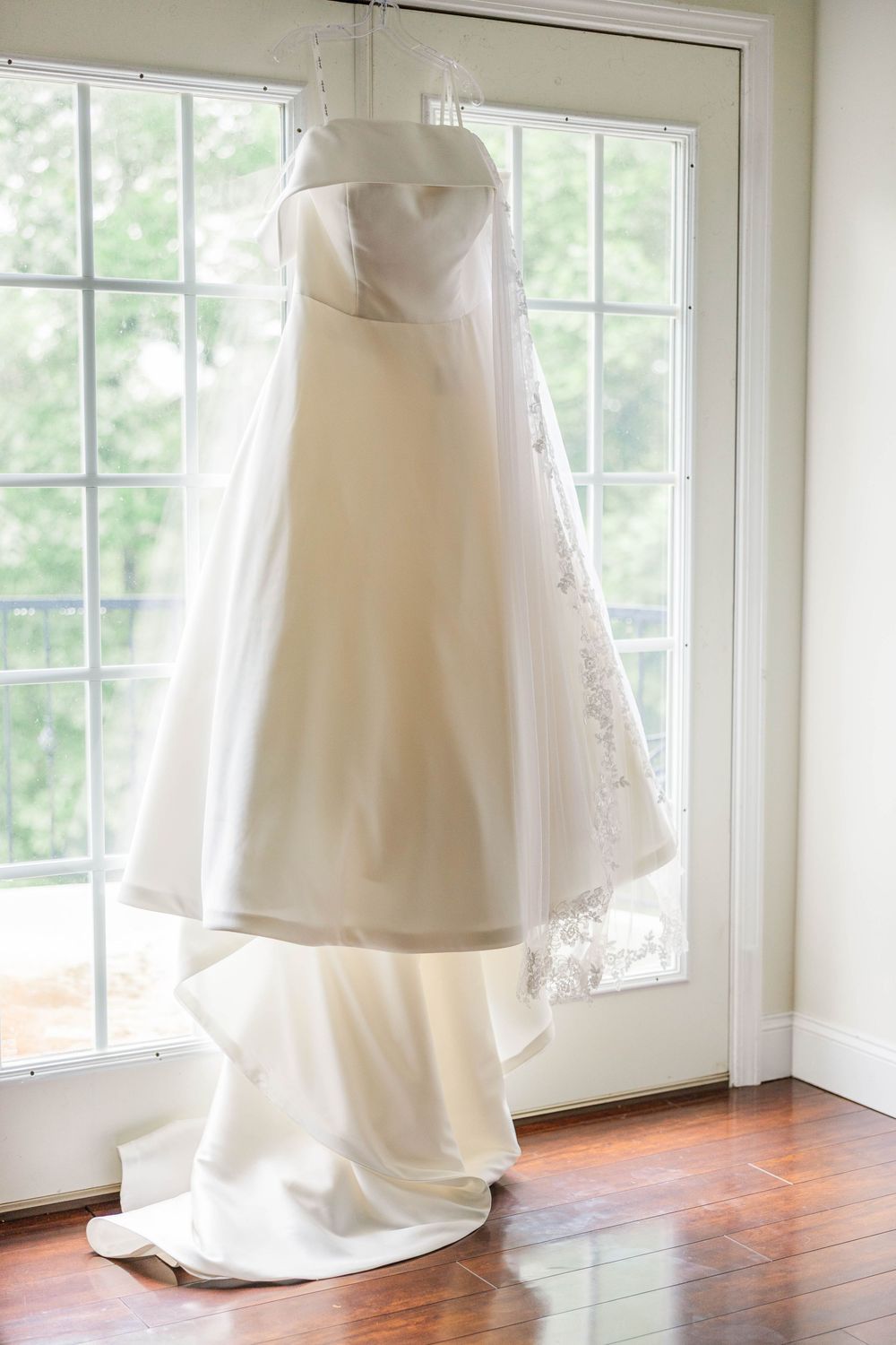 Elegant white wedding dress hanging in sunlit room with hardwood floors and french doors.
