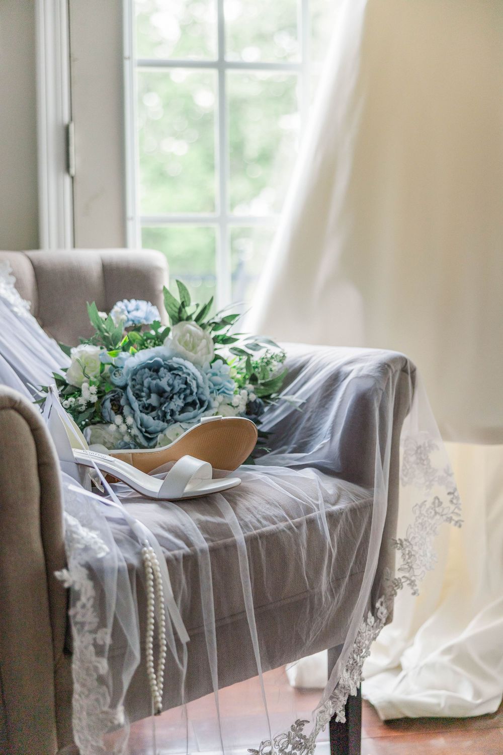 Wedding bouquet with blue and white flowers rests on a grey chair near a sunlit window with flowing curtains.