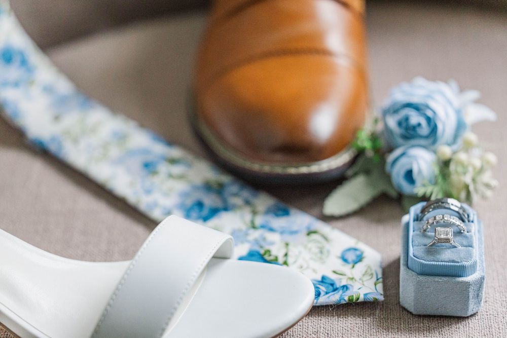 White bridal sandals and brown leather shoes displayed with blue floral details and a velvet ring box.