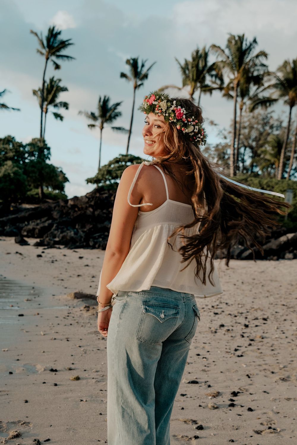 Person wearing floral crown and white tank top walks on tropical beach with palm trees and volcanic rocks in Hawaii.