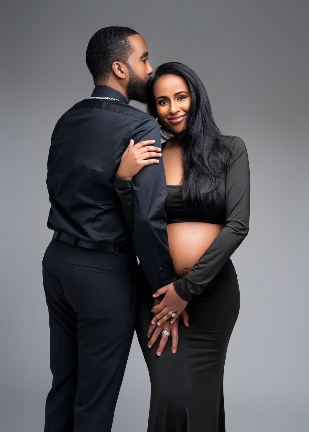An expecting couple wearing all black embraces in an intimate maternity photoshoot against a gray background