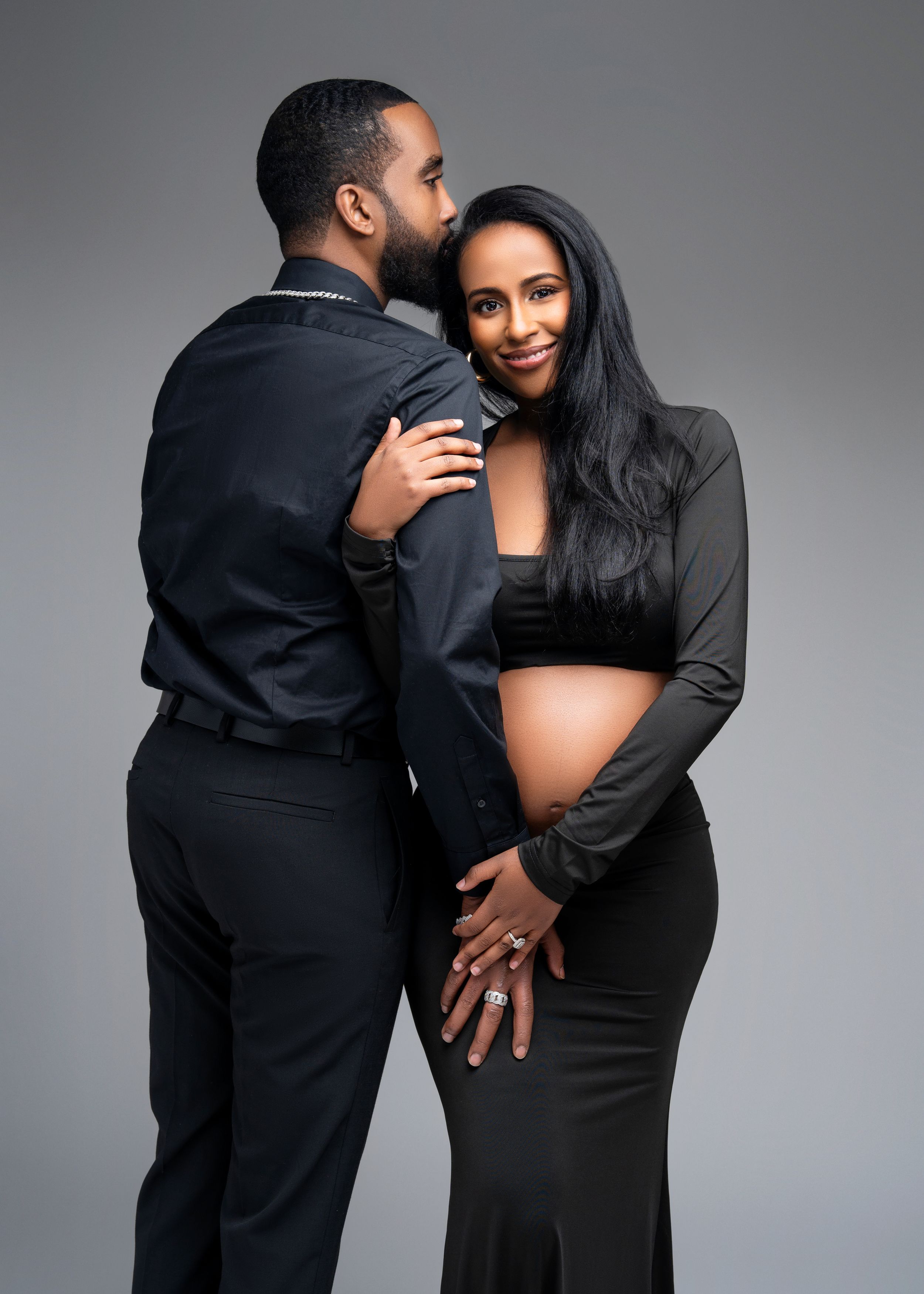 An expecting couple wearing all black embraces in an intimate maternity photoshoot against a gray background