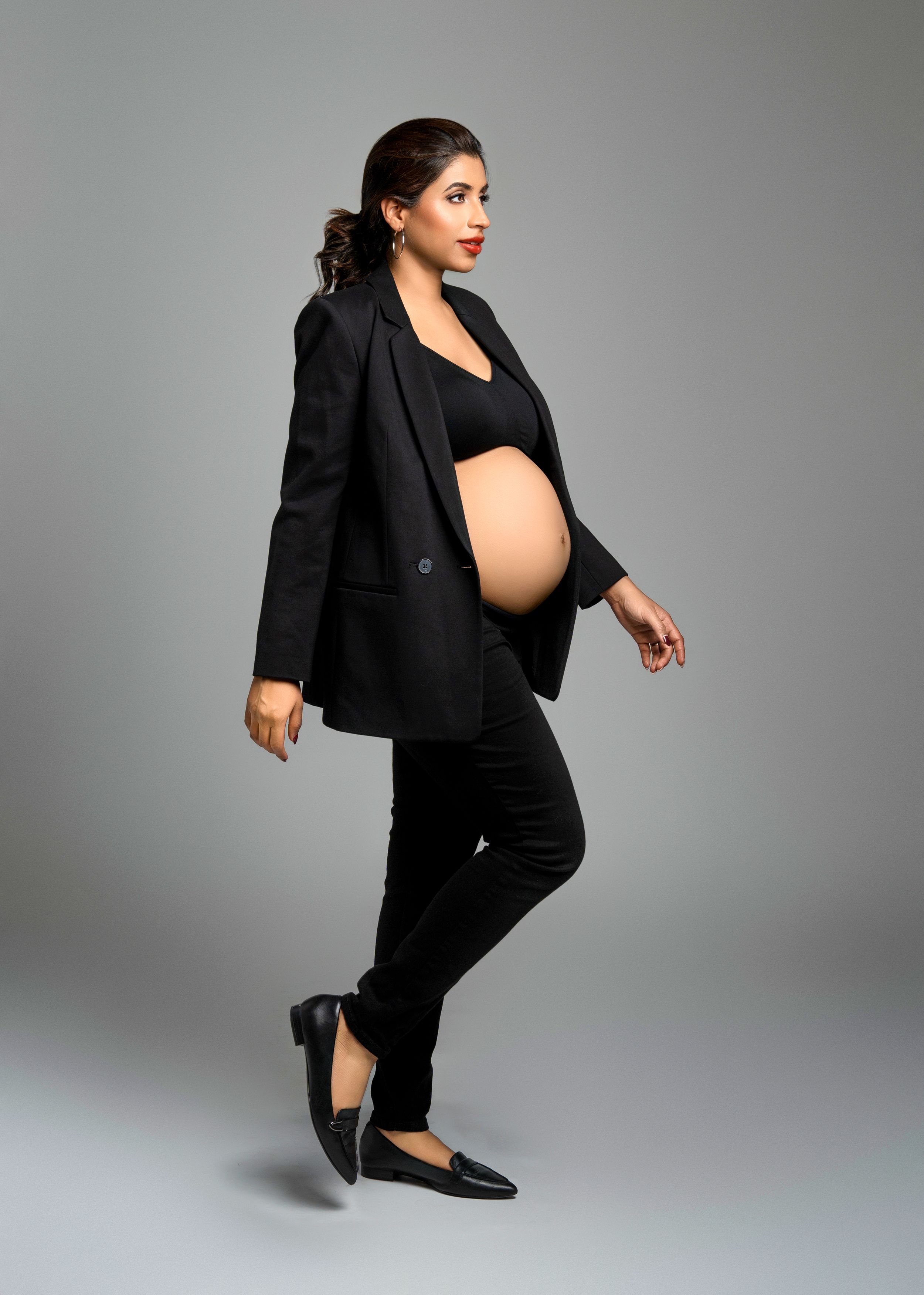 Stylish pregnant woman in black business attire and flats strikes a confident pose against gray studio background.