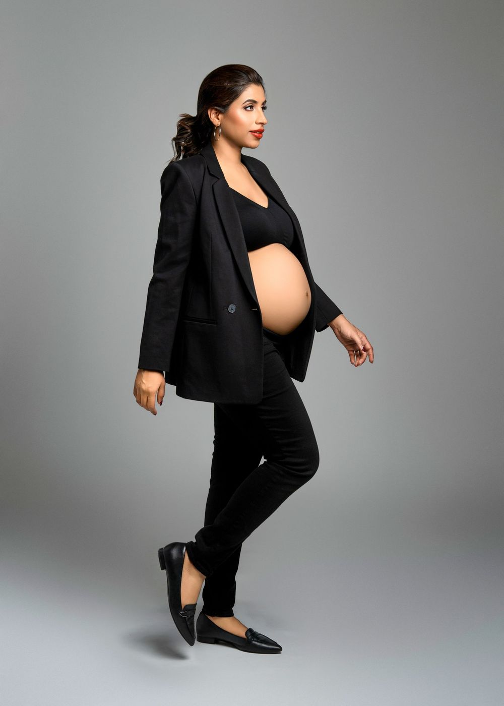 Stylish pregnant woman in black business attire and flats strikes a confident pose against gray studio background.