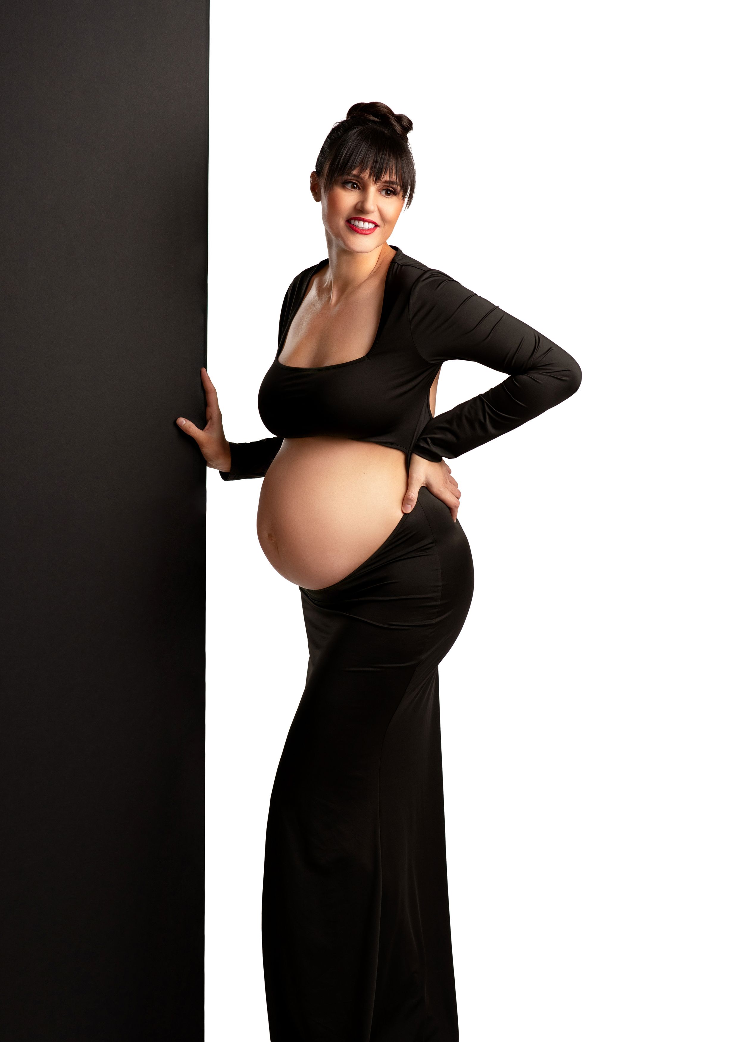 Maternity portrait of an expecting mother in a black two piece gown posing against a dark and light backdrop.