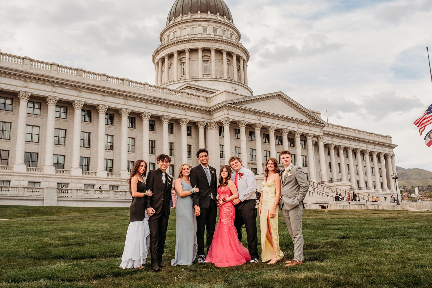 A group of elegantly dressed people pose for a photo in front of the Utah State Capitol building on a cloudy day.