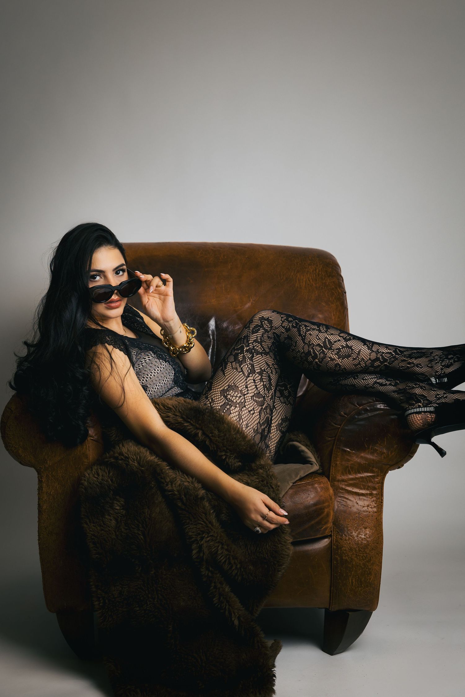 Woman in lace outfit lounging on brown leather armchair in moody boudoir photoshoot.