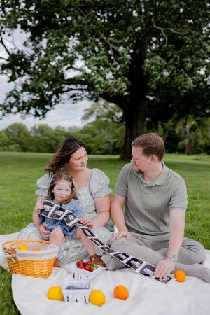 Family picnicking on a white blanket with oranges and snacks under a leafy tree.