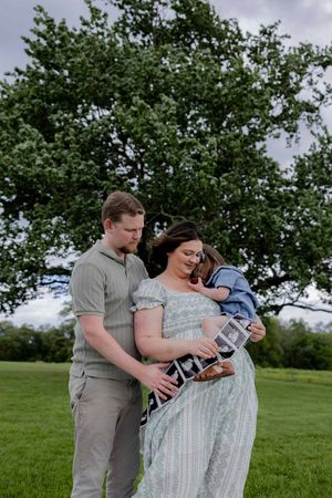 A tender family moment outdoors under a leafy tree with a small child and expecting parents.