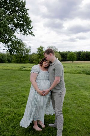 A romantic couple embraces on a lush green field under cloudy skies, with her wearing a flowing pale blue dress.