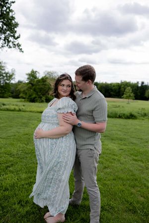 An expectant couple embraces on a green lawn under cloudy skies, her wearing a flowy dress.