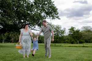 A family of three plays together on a grassy field during a cloudy day, with the child in a blue outfit.