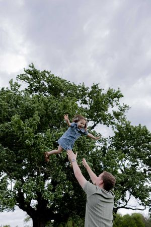 A playful moment as a parent tosses a small child high in the air against a backdrop of trees and cloudy sky.